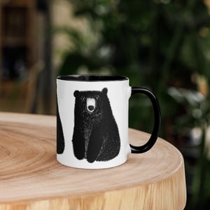 May include: A white ceramic mug with a black rim and handle. The mug features a black and white illustration of a bear.