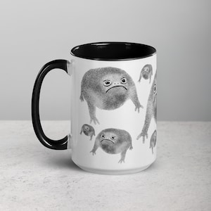 May include: White ceramic coffee mug with a black rim and handle. The mug features a repeating pattern of black and white illustrations of grumpy frogs.