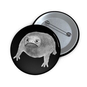 May include: A black and white button featuring a cartoon frog with a grumpy expression. The frog is depicted in a simple, minimalist style.