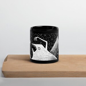 Pode incluir: Caneca de cer&acirc;mica preta com uma ilustra&ccedil;&atilde;o branca de uma criatura com dentes e garras afiadas, em um fundo de noite estrelada. O design &eacute; art&iacute;stico, ideal para bebidas quentes.