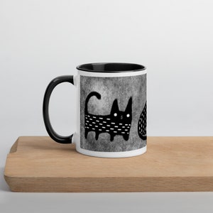 May include: White ceramic mug with a black rim and handle. The mug features a black and white illustration of two cats on a gray background.