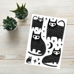 May include: A sheet of black and white stickers featuring four stylised black cats with skull details. The cats are surrounded by small stars and a crescent moon.