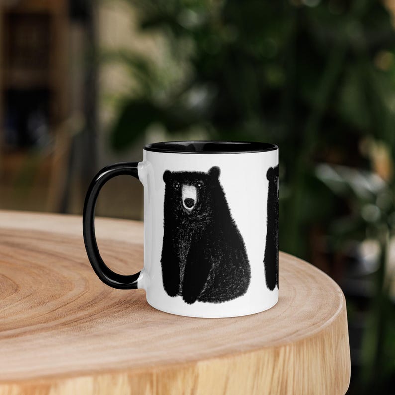May include: White ceramic mug with a black rim and handle. The mug features a black and white illustration of a bear sitting on a white background.