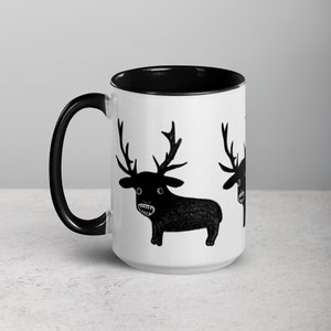 May include: White ceramic mug with a black rim and handle. The mug features a black and white illustration of a cartoon deer with large eyes and a wide open mouth. The deer is repeated on the mug.
