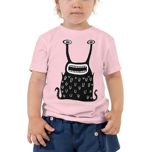 May include: A pink toddler t-shirt with a black and white cartoon monster graphic. The monster has a large mouth with teeth and a series of U-shaped markings on its body.