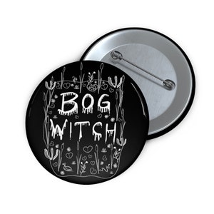 May include: A black button with the words "BOG WITCH" in white, surrounded by a white botanical design. The button is round and has a pin on the back. The second button is visible in the background.