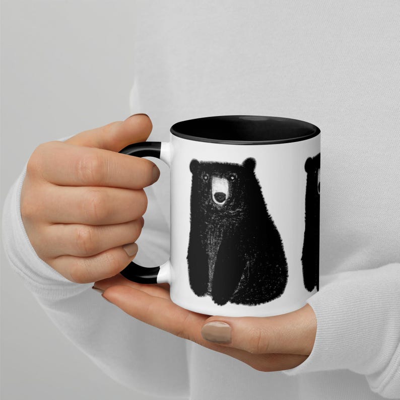 May include: A white ceramic mug with a black rim and handle. The mug features a black and white illustration of a bear sitting on a white background.