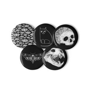 May include: Set of five black and white button badges featuring various designs. The designs include a skull, a cat, a moth, an eye pattern, and a skull with teeth.