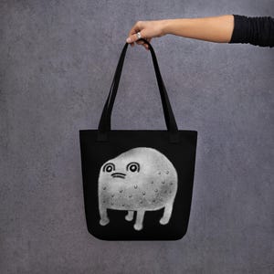 May include: Black tote bag featuring a white graphic of a round, furry creature with large eyes and a small mouth. The creature has four short legs and is standing on a black background.