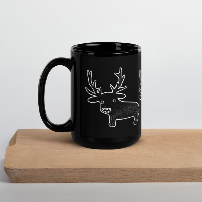 May include: Black ceramic mug featuring a white cartoon deer illustration. The deer has large antlers and a toothy grin. The mug is sitting on a wooden surface.