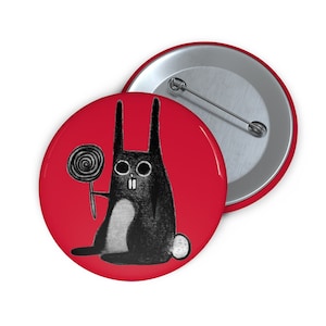 Puede incluir: Un botón rojo con una ilustración en blanco y negro de un conejo sosteniendo un caramelo. El conejo tiene orejas largas y una barriga blanca. Se ve un segundo botón con un alfiler plateado.
