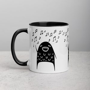 May include: A white ceramic mug with a black rim and handle. The mug features a black and white illustration of a singing monster with musical notes around it.