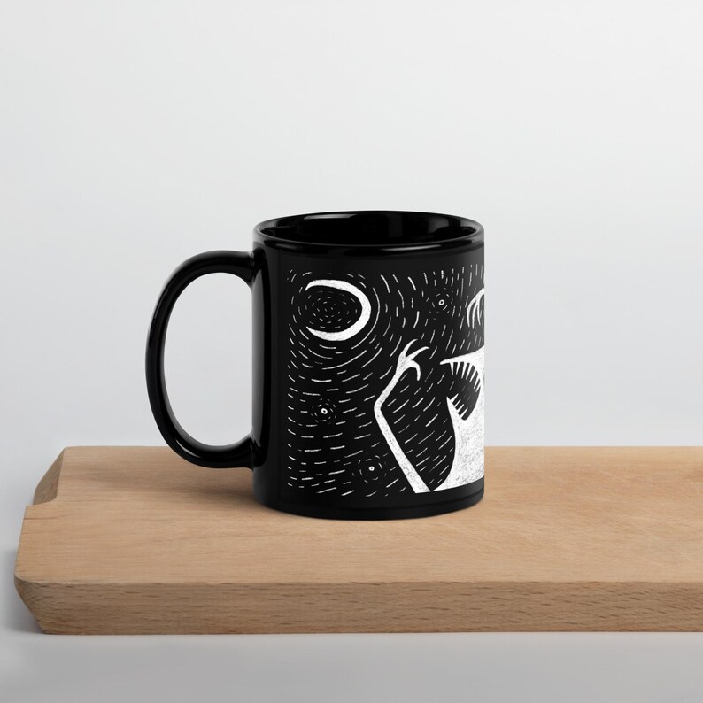 Pode incluir: Caneca de cer&acirc;mica preta com uma ilustra&ccedil;&atilde;o branca de uma criatura sob uma lua crescente e estrelas. A caneca tem uma al&ccedil;a curva e um acabamento brilhante. O design &eacute; de estilo gr&aacute;fico minimalista.