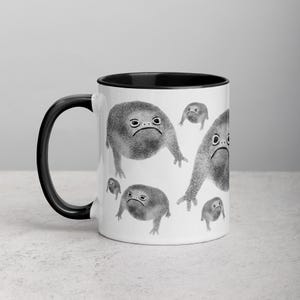 以下が含まれることがあります： 黒色の縁取りと取っ手のある白いセラミック製のコーヒーカップ。カップには、不機嫌そうな表情をした白黒の漫画のカエルが繰り返し描かれています。
