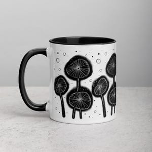 May include: A white ceramic mug with a black rim and handle. The mug features a black and white illustration of stylized dandelion-like flowers with a speckled background.