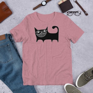 K&ouml;nnte beinhalten: Ein pinkfarbenes T-Shirt mit einer schwarz-wei&szlig;en Illustration einer Katze mit einem breiten L&auml;cheln. Die Katze hat gro&szlig;e Augen und einen langen Schwanz.