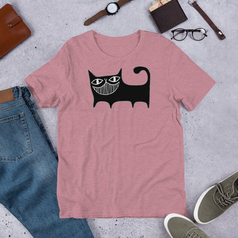 K&ouml;nnte beinhalten: Ein pinkfarbenes T-Shirt mit einer schwarz-wei&szlig;en Illustration einer Katze mit einem breiten L&auml;cheln. Die Katze hat gro&szlig;e Augen und einen langen Schwanz.