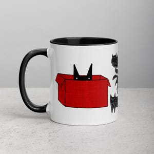 May include: A white ceramic mug with a black rim and handle. The mug features a black cat peeking out of a red box with other black cats on the side.