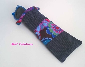 May include: A fabric glasses case featuring a dark gray ribbed textile top and bottom section with a central band of floral fabric in shades of blue, pink, and green. The case contains blue sunglasses and displays the text @n7 Créations.