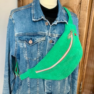 Puede incluir: Un riñonera de pana verde brillante con una cremallera roja y blanca. La bolsa se lleva sobre una chaqueta vaquera y una camiseta negra. La riñonera tiene un tirador de cremallera plateado.