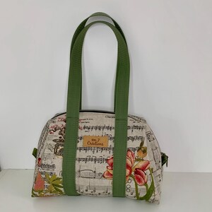 May include: A beige fabric handbag with a musical score pattern and floral accents. The bag has green handles and a zipper closure. A small brown tag reads "on 7 Créations".