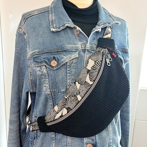 May include: A crossbody bag featuring a black corduroy section and a cream-colored panel with a black dotted floral pattern. The bag has a silver zipper with two pull tabs, a small red accent detail, and an adjustable strap worn over a denim jacket.