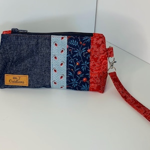 Peut inclure: Une pochette zippée en denim bleu et tissu floral avec une sangle rouge et une étiquette en cuir marron qui dit "on 7 Creations".