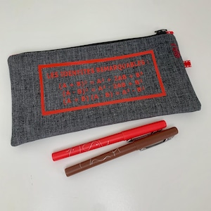 May include: A gray pencil case with a black zipper and red accents. The case features red text that reads "LES IDENTITES REMARQUABLES" and mathematical formulas. Two pens, one red and one brown, are lying in front of the case.