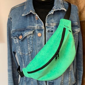 Peut inclure: Une banane en velours côtelé vert vif avec une fermeture éclair et une sangle noires. La banane est portée sur une veste en jean.