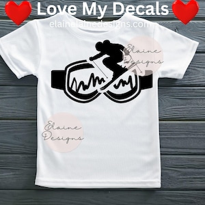 May include: White t-shirt with a black silhouette of a skier skiing over goggles. The goggles have a wavy design inside. The text "Love My Decals" and "elainedesigns.com" are at the top. The design is suitable for winter sports enthusiasts.