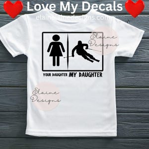 May include: White t-shirt with a black graphic. The graphic shows a stick figure of a woman and a skier, with the text "YOUR DAUGHTER MY DAUGHTER" below. The shirt also has the text "Love My Decals" at the top.