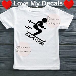 May include: White t-shirt featuring a black skier graphic with the text "ELAINE ELAINE" and "Love My Decals". The design includes a skier in motion with ski poles and speed lines. The shirt also has the text "elainedesigns.com".