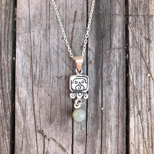 May include: Silver necklace with a square pendant featuring a Mayan glyph and a green stone bead.
