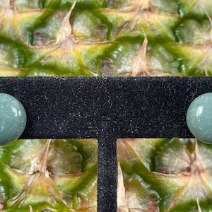 Op de afbeelding: Een paar ronde, lichtgroene oorstekers. Elke oorbel is een gladde bol met een glanzende afwerking. De oorbellen worden gepresenteerd op een zwart fluwelen oppervlak tegen een achtergrond van een ananas.