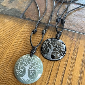 Puede incluir: Dos colgantes de árbol de la vida en cordones negros. Un colgante es negro con un diseño de árbol plateado, el otro es verde con un diseño de árbol blanco.