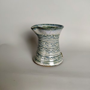 May include: A small, white ceramic jug with a blue and green glaze. The jug has a decorative pattern around the body.
