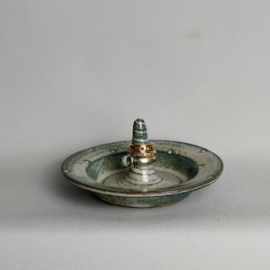 May include: A ceramic ring holder with a green and white striped design. The holder has a small dish and a raised centre post for rings.