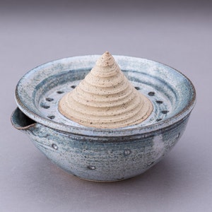May include: A ceramic citrus juicer with a light blue bowl and a central reamer. The bowl has a spout and drainage holes. The reamer is cone-shaped and unglazed, with a textured surface. The juicer is designed for kitchen use.