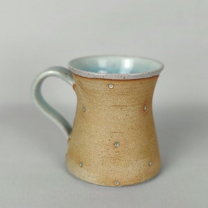 May include: A light brown ceramic mug with a light blue interior and white speckles. The mug has a rounded body and a handle.