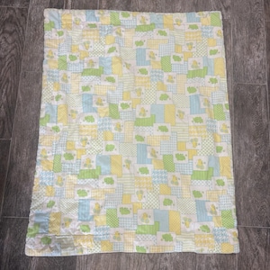 May include: A rectangular baby quilt featuring a patchwork design of light blue, pale yellow, and lime green squares. The fabric displays repeating patterns of small green frogs and yellow ducks, interspersed with geometric textures like dots and stripes.