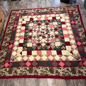May include: A square patchwork quilt featuring a geometric pattern of diamonds and triangles in shades of cream, pink, burgundy, and dark green. The quilt is bordered by a wide band of fabric with a floral and leaf print in muted earth tones.