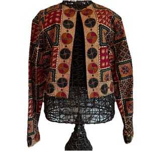 May include: A patterned jacket with a unique design. The jacket features a mix of geometric shapes and circular patterns in shades of red, green, brown, and beige. The jacket is displayed on a wire mannequin.