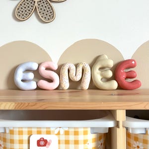 Op de afbeelding: Een kleurrijke naamweergave met de naam "ESMEE" in grote, afgeronde letters. De letters zijn in tinten wit, roze, gespikkeld, crème en rood. De display staat op een houten plank, met opbergbakken eronder.