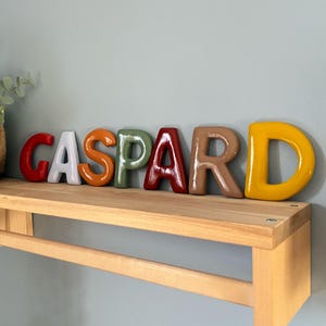 Könnte beinhalten: Der Name "Gaspard" wird durch farbige Keramikbuchstaben auf einem Holzregal dargestellt. Die Buchstaben sind rot, weiß, orange, grün, weinrot, beige und gelb. Das Regal ist an einer hellblauen Wand befestigt.