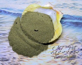 May include: A large, off-white scallop shell filled with a mound of fine, olive-green powder. The shell is positioned on a surface with a printed ocean wave pattern. The text MT Pet Emporium is visible in the lower right corner of the image.