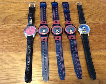 May include: Five Mickey Mouse watches with different designs. Three watches have black and blue bands with red stars. One watch has a black leather band with a red face. One watch has a black leather band with a gold face.