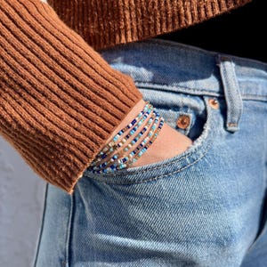 May include: A stack of beaded bracelets in shades of blue, white, and gold. The bracelets are worn on a wrist, with a brown sweater sleeve and blue jeans in the background. The bracelets are made of small, colorful beads.
