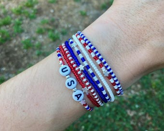 May include: A collection of beaded bracelets in red, white, and blue, with a patriotic theme. One bracelet spells out "USA" with letter beads. The bracelets are adorned with small star charms, perfect for celebrating Independence Day.