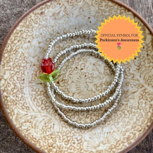 May include: A beaded bracelet with silver beads and a red and green flower accent. The bracelet is displayed on a decorative plate. The image includes a text graphic that reads "OFFICIAL SYMBOL FOR Parkinson's Awareness".