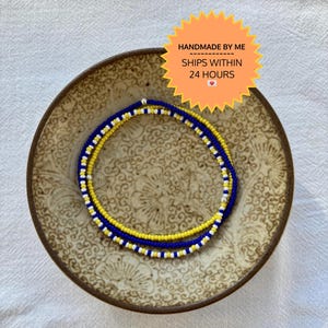 May include: Three beaded bracelets in blue, yellow, and white, resting on a decorative brown plate. The bracelets are made of small, colorful beads. The plate has a floral pattern. Text on the image reads "HANDMADE BY ME SHIPS WITHIN 24 HOURS".
